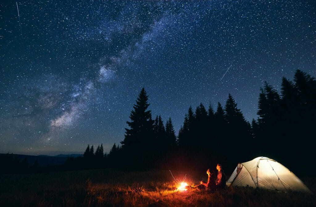 The milky way shines above a forest where two people sit next to a tent and campfire.