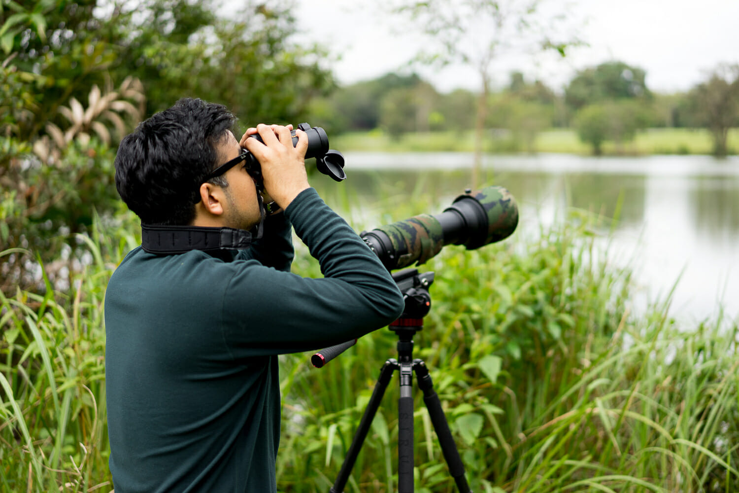 A photographer using binoculars with a camera mounted on a tripod by a lakeside.