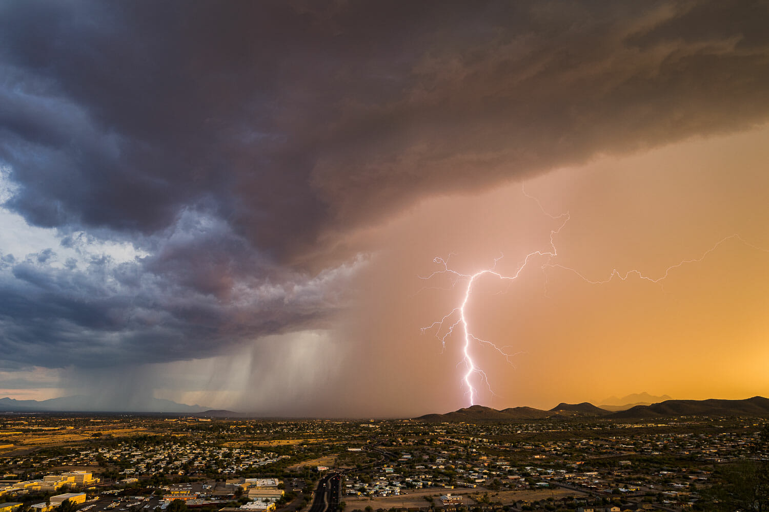 Lightning strike beneath storm clouds over a cityscape at sunset.