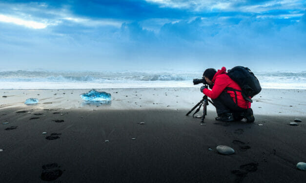 Photographer capturing an image of ice on a black sand beach under a cloudy sky.