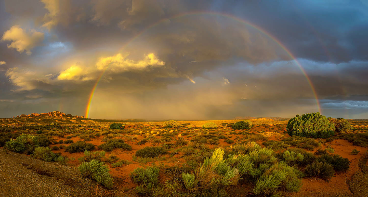 A panoramic view of a full rainbow over a desert landscape at sunset.