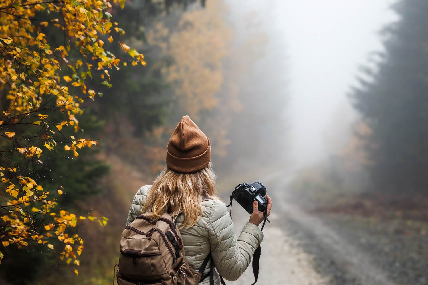 Woman with camera standing on a foggy forest road surrounded by autumn foliage.