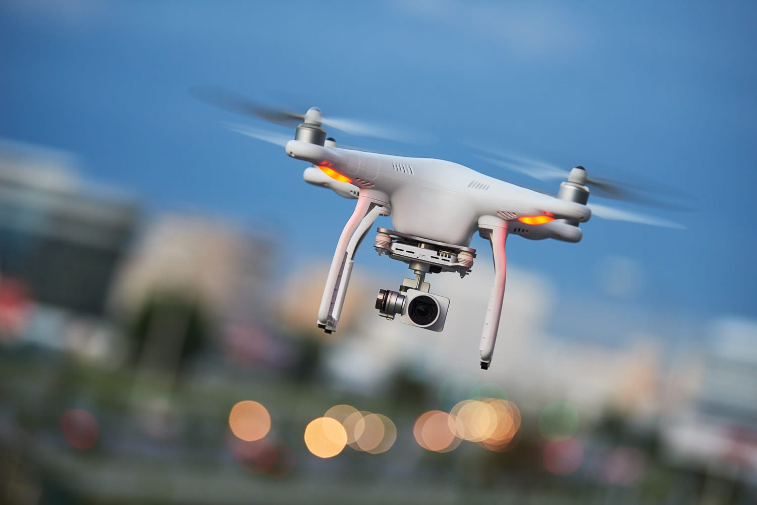 Quadcopter drone with camera flying against a blurred cityscape background.