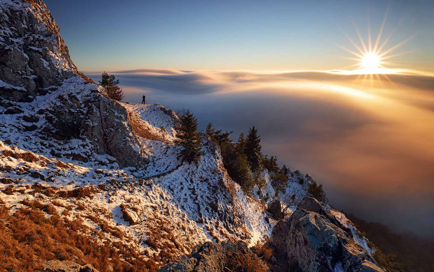 Hiker standing on a snowy mountain trail at sunset, with sea of clouds below.