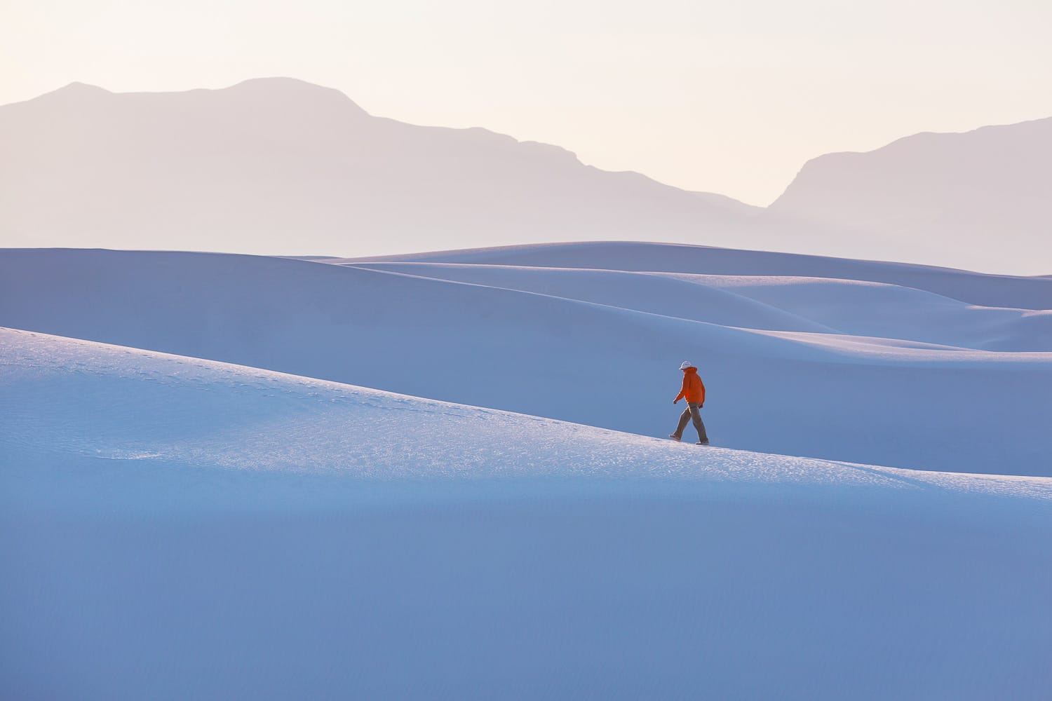 A lone hiker walks across smooth sand dunes with mountain silhouettes in the background during a hazy sunrise.