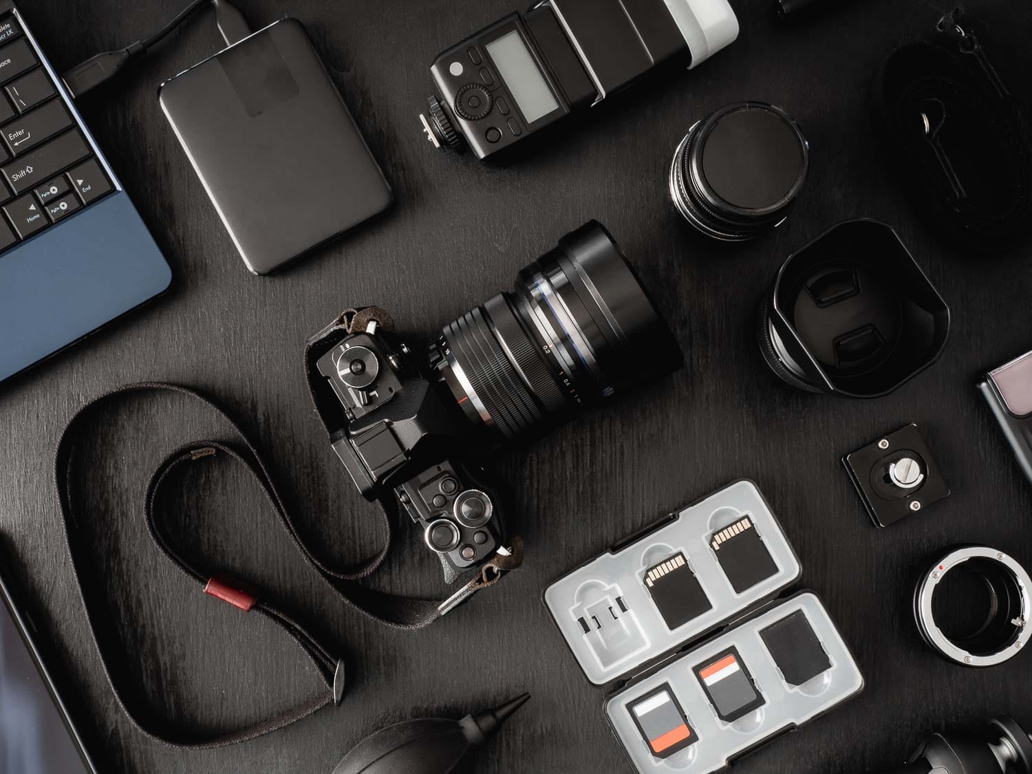 Assorted photography equipment arranged on a dark surface, including a camera, lenses, and memory cards.